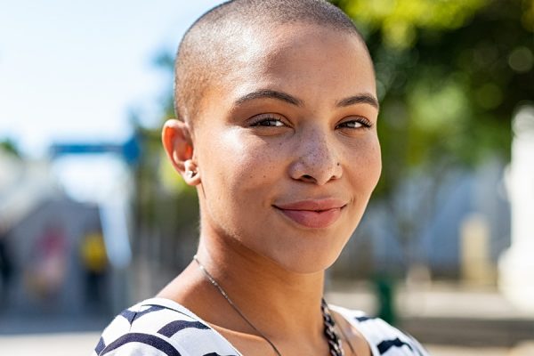 Portrait of young happy bald woman on city street looking at camera. Confident stylish girl outdoor with copy space. Proud and satisfied black curvy woman standing on street.