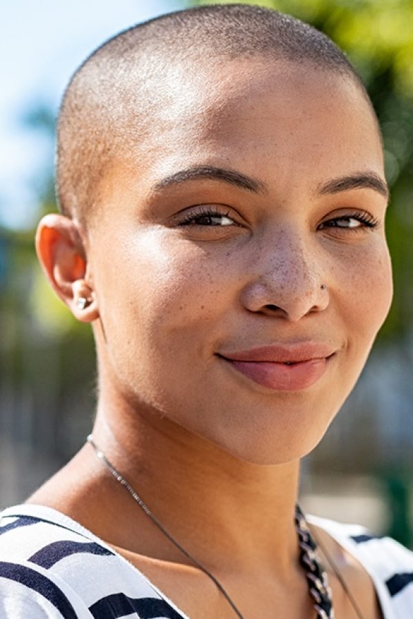 Portrait of young happy bald woman on city street looking at camera. Confident stylish girl outdoor with copy space. Proud and satisfied black curvy woman standing on street.
